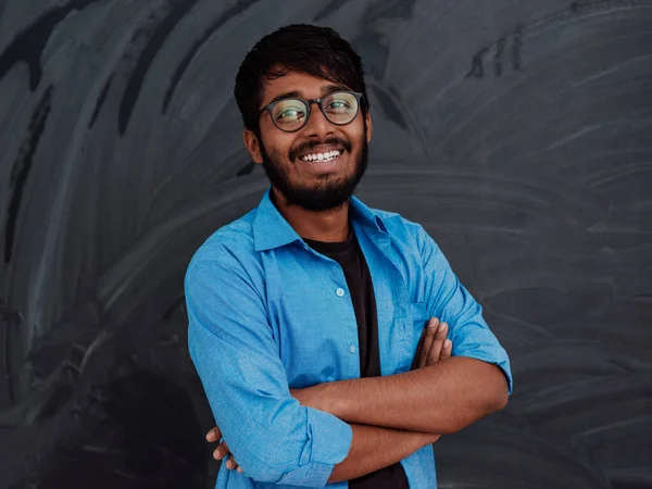 一名身穿蓝色衬衫 双手举着眼镜的印度青年学生在学校黑板前交叉站立 — 图库照片