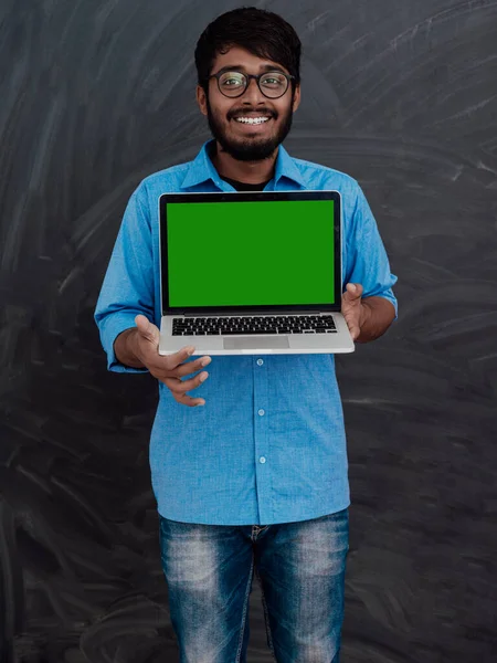 stock image Indian smiling young student in blue shirt and glasses using a laptop and posing on school blackboard background. 