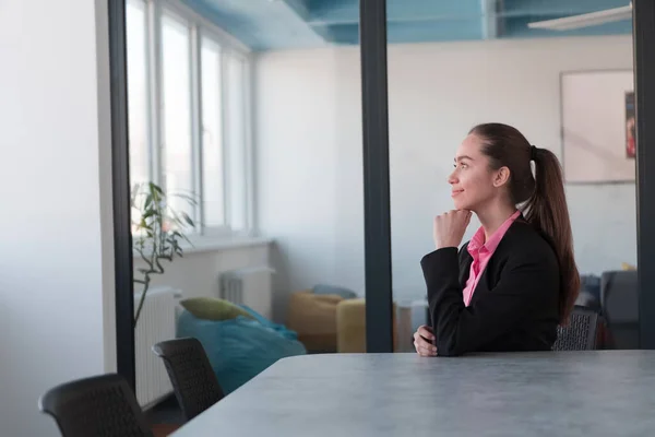 成功的年轻女性领导者 身穿一件粉色衬衫 坐在现代玻璃办公室里 脸上带着坚定的微笑 — 图库照片