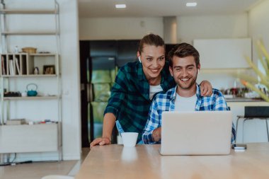 A young married couple is talking to parents, family and friends on a video call via a laptop while sitting in the living room of their modern house. clipart