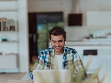 Modern bir oturma odasındaki masada oturan adam, kulaklıklarıyla iş görüşmesi için dizüstü bilgisayar kullanıyor, arkadaşlarıyla sohbet ediyor ve eğleniyor..
