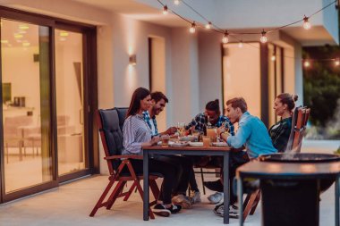 Bir grup genç insan akşamları modern bir evin terasında yemek yiyorlar. Dostlar ve aile için eğlence. Tatil kutlamaları, barbekülü düğünler.