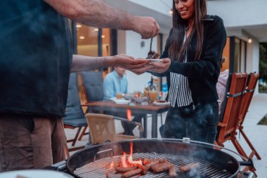 Bir grup arkadaş ve aile akşamları terasta büyük, modern bir evin önünde barbekü yapıyorlar..