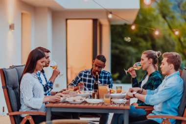 Bir grup genç insan akşamları modern bir evin terasında yemek yiyorlar. Dostlar ve aile için eğlence. Tatil kutlamaları, barbekülü düğünler.