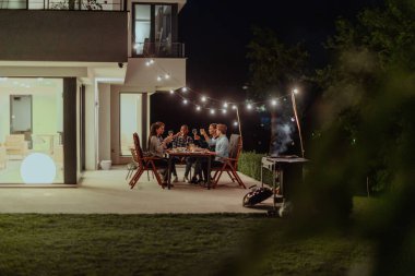 Bir grup genç insan akşamları modern bir evin terasında yemek yiyorlar. Dostlar ve aile için eğlence. Tatil kutlamaları, barbekülü düğünler.
