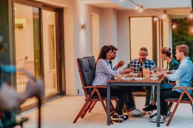 Bir grup genç insan akşamları modern bir evin terasında yemek yiyorlar. Dostlar ve aile için eğlence. Tatil kutlamaları, barbekülü düğünler.