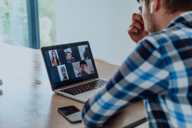 Modern bir oturma odasındaki masada oturan adam, kulaklıklarıyla iş görüşmesi için dizüstü bilgisayar kullanıyor, arkadaşlarıyla sohbet ediyor ve eğleniyor..