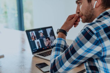 Modern bir oturma odasındaki masada oturan adam, kulaklıklarıyla iş görüşmesi için dizüstü bilgisayar kullanıyor, arkadaşlarıyla sohbet ediyor ve eğleniyor..