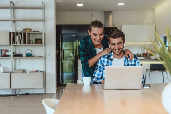 A young married couple is talking to parents, family and friends on a video call via a laptop while sitting in the living room of their modern house.