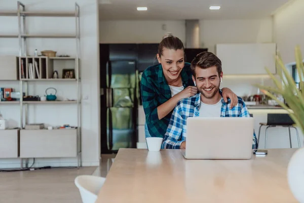 A young married couple is talking to parents, family and friends on a video call via a laptop while sitting in the living room of their modern house.
