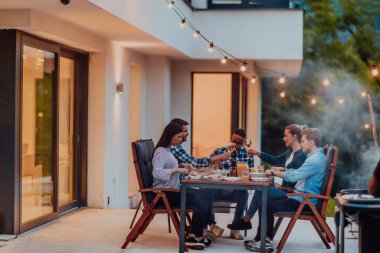 Bir grup genç insan akşamları modern bir evin terasında yemek yiyorlar. Dostlar ve aile için eğlence. Tatil kutlamaları, barbekülü düğünler.