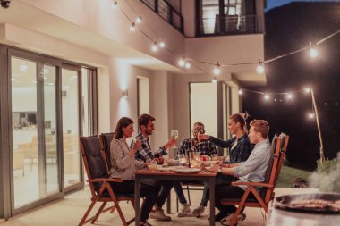 Bir grup genç insan akşamları modern bir evin terasında yemek yiyorlar. Dostlar ve aile için eğlence. Tatil kutlamaları, barbekülü düğünler.