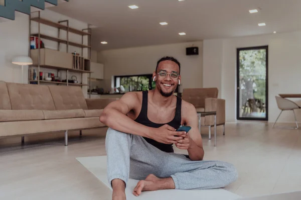 アフリカ系アメリカ人男性の頭部を眼鏡で撮影し スマートフォンでヘッドフォンをし リビングルームの床に座って — ストック写真