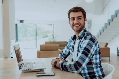 Modern bir oturma odasındaki masada oturan adam, kulaklıklarıyla iş görüşmesi için dizüstü bilgisayar kullanıyor, arkadaşlarıyla sohbet ediyor ve eğleniyor..