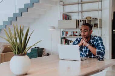 Modern bir oturma odasındaki masada oturan gözlüklü Afrikalı Amerikalı bir adam bilgisayarını iş videosu sohbeti, arkadaşlarla sohbet ve eğlence için kullanıyor..