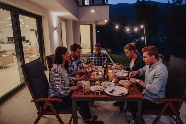A group of young diverse people having dinner on the terrace of a modern house in the evening. Fun for friends and family. Celebration of holidays, weddings with barbecue