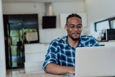 Modern bir oturma odasındaki masada oturan gözlüklü Afrikalı Amerikalı bir adam bilgisayarını iş videosu sohbeti, arkadaşlarla sohbet ve eğlence için kullanıyor..