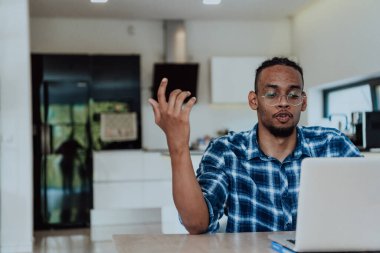 Modern bir oturma odasındaki masada oturan gözlüklü Afrikalı Amerikalı bir adam bilgisayarını iş videosu sohbeti, arkadaşlarla sohbet ve eğlence için kullanıyor..