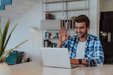 Modern bir oturma odasındaki masada oturan adam, kulaklıklarıyla iş görüşmesi için dizüstü bilgisayar kullanıyor, arkadaşlarıyla sohbet ediyor ve eğleniyor..