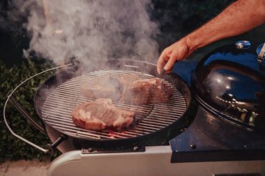 Lezzetli etin ızgarada pişirildiği yakın plan fotoğraf. Arka planda, arkadaşlar ve aile oturup yemek bekliyor..