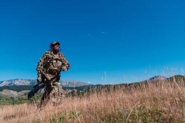Ordu askeri dürbünlü bir keskin nişancı tüfeği tutuyor ve ormanda yürüyor. savaş, ordu, teknoloji ve insanlar kavramı