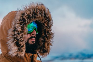 Soğuk ve karlı bir bölgede, kalın kahverengi bir ceketli, kar gözlüklü ve eldivenli bir adamın vesikalık fotoğrafı. Ülkenin soğuk bölgelerinde yaşam