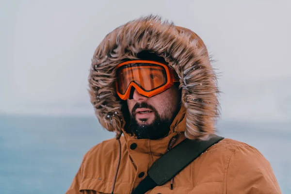 Headshot photo of a man in a cold snowy area wearing a thick brown winter jacket, snow goggles and gloves. Life in cold regions of the country