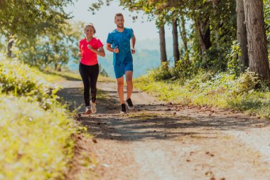 Couple enjoying in a healthy lifestyle while jogging on a country road through the beautiful sunny forest, exercise and fitness concept. 
