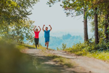 Couple enjoying in a healthy lifestyle while jogging on a country road through the beautiful sunny forest, exercise and fitness concept. 