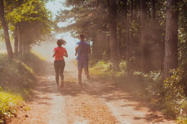 Couple enjoying in a healthy lifestyle while jogging on a country road through the beautiful sunny forest, exercise and fitness concept. 