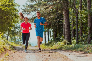 Couple enjoying in a healthy lifestyle while jogging on a country road through the beautiful sunny forest, exercise and fitness concept. 