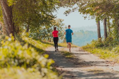 Couple enjoying in a healthy lifestyle while jogging on a country road through the beautiful sunny forest, exercise and fitness concept. 