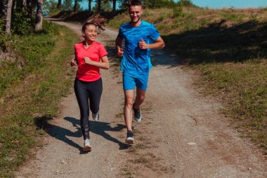 Couple enjoying in a healthy lifestyle while jogging on a country road through the beautiful sunny forest, exercise and fitness concept. 