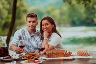 Happy couple toasting red wine glass while having picnic french dinner party outdoor during summer holiday vacation near the river at beautiful nature.
