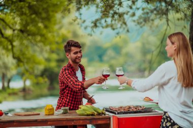 Happy couple toasting red wine glass while having picnic french dinner party outdoor during summer holiday vacation near the river at beautiful nature.