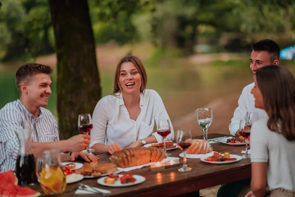 Group of happy friends having picnic french dinner party outdoor during summer holiday vacation near the river at beautiful nature.