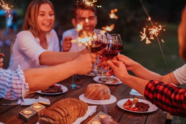 Group of happy friends celebrating holiday vacation using sprinklers and drinking red wine while having picnic french dinner party outdoor near the river on beautiful summer evening in nature.