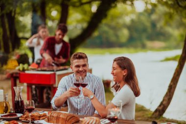 Happy couple toasting red wine glass while having picnic french dinner party outdoor during summer holiday vacation near the river at beautiful nature.