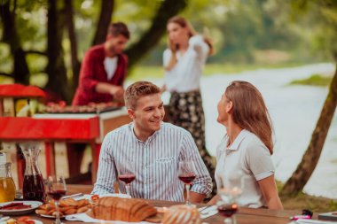 Happy couple toasting red wine glass while having picnic french dinner party outdoor during summer holiday vacation near the river at beautiful nature.