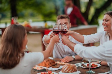 Bir grup mutlu arkadaş kırmızı şarap kadehi içerken dışarıda piknik yapıyorlar yaz tatili boyunca nehir kenarında güzel doğada..