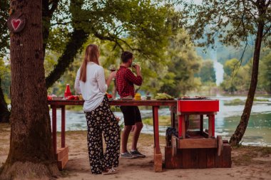Happy couple toasting red wine glass while having picnic french dinner party outdoor during summer holiday vacation near the river at beautiful nature.