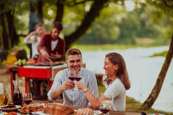 Happy couple toasting red wine glass while having picnic french dinner party outdoor during summer holiday vacation near the river at beautiful nature.