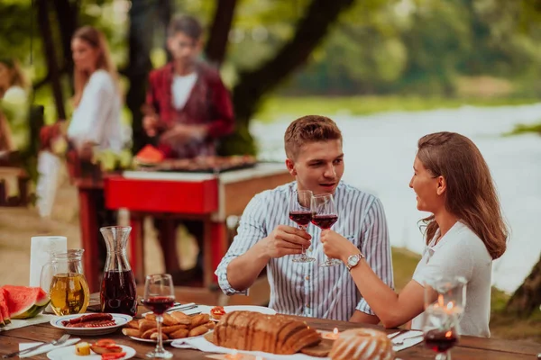 Happy couple toasting red wine glass while having picnic french dinner party outdoor during summer holiday vacation near the river at beautiful nature.