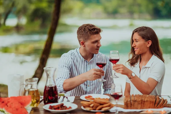 Happy couple toasting red wine glass while having picnic french dinner party outdoor during summer holiday vacation near the river at beautiful nature.