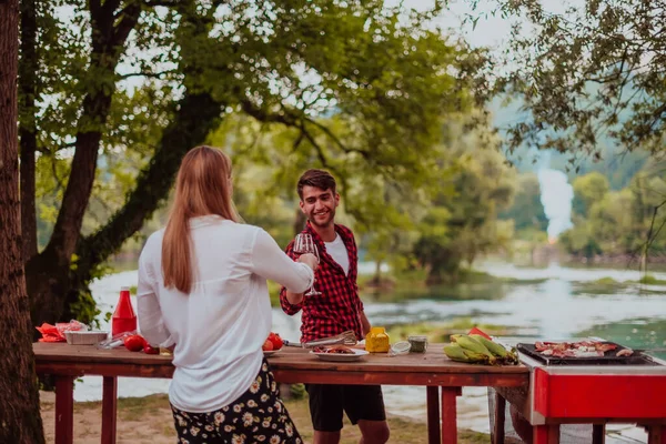 Happy couple toasting red wine glass while having picnic french dinner party outdoor during summer holiday vacation near the river at beautiful nature.