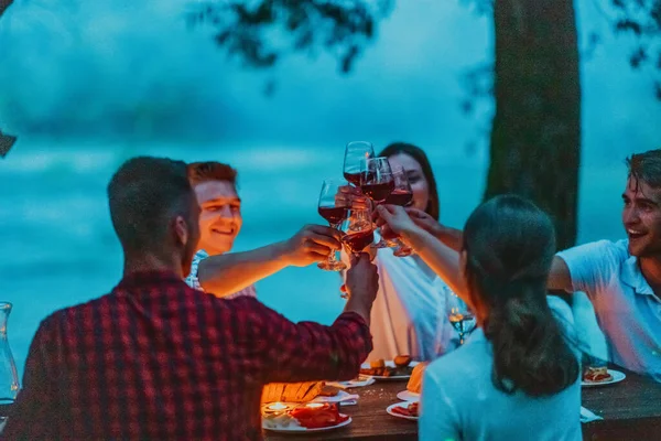 Group of happy friends celebrating holiday vacation using sprinklers and drinking red wine while having picnic french dinner party outdoor near the river on beautiful summer evening in nature.