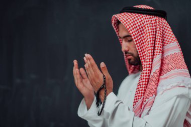 Arabian man in traditional clothes making traditional prayer to God, keeps hands in praying gesture in front of black chalkboard representing modern islam fashion and ramadan kareem concept. 