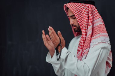 Arabian man in traditional clothes making traditional prayer to God, keeps hands in praying gesture in front of black chalkboard representing modern islam fashion and ramadan kareem concept. 