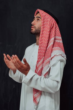Arabian man in traditional clothes making traditional prayer to God, keeps hands in praying gesture in front of black chalkboard representing modern islam fashion and ramadan kareem concept. 