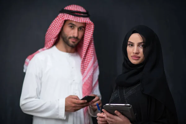 若いイスラム教徒のビジネスカップルアラビア人男性と女性でファッショナブルなヒジャーブドレスを使用して携帯電話とタブレットコンピュータ黒の黒板の前に現代のイスラム教徒のファッション技術を表す — ストック写真